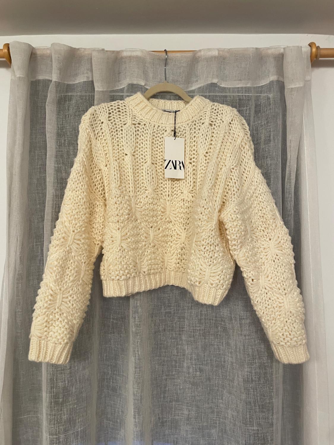 Cream-colored chunky knit sweater hanging on a hanger in front of a gray curtain. Visible label with the text "ZARA".