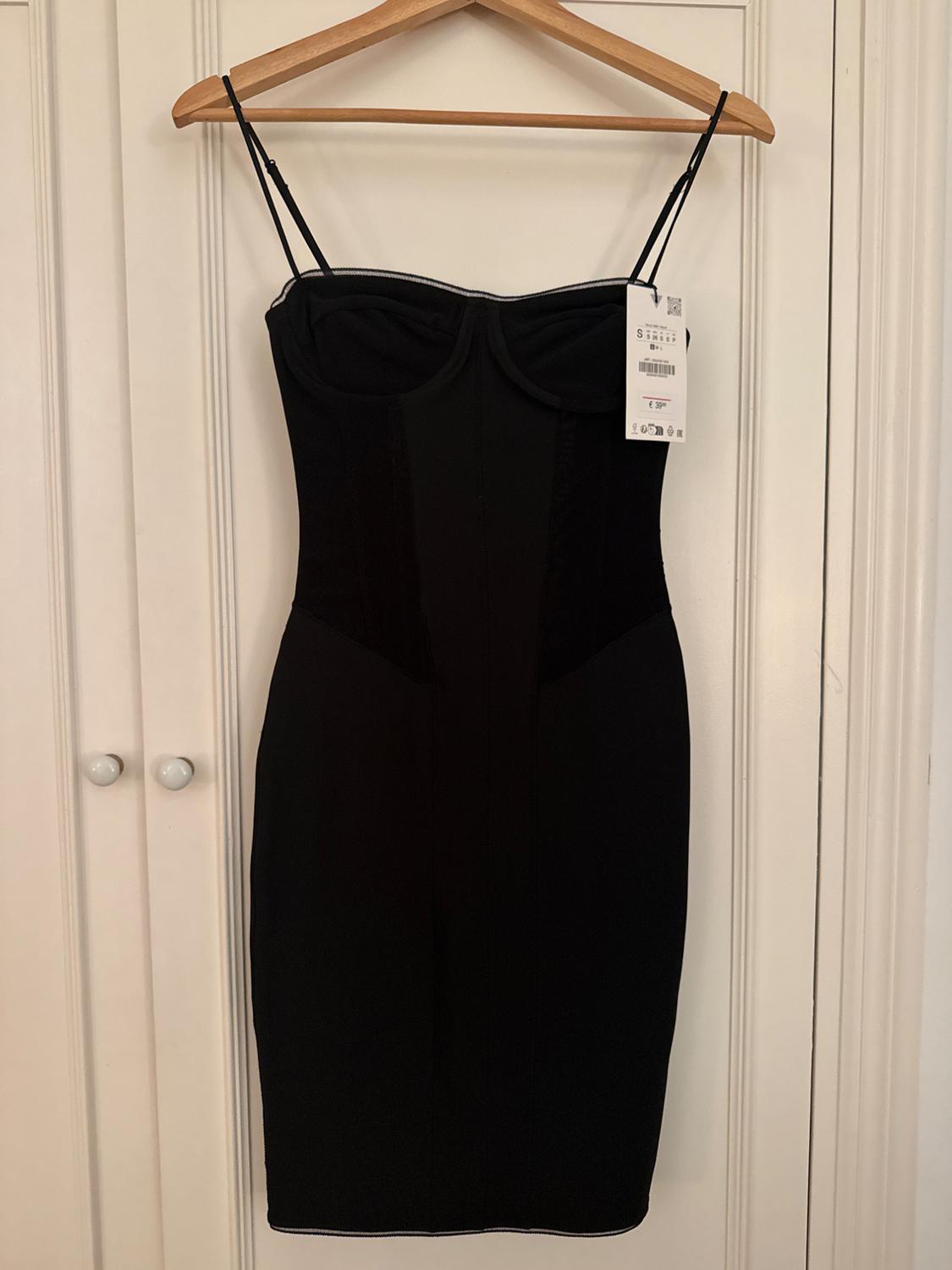 Tight black dress with spaghetti straps, hanging on a wooden hanger. Visible label on the right side. White door background.