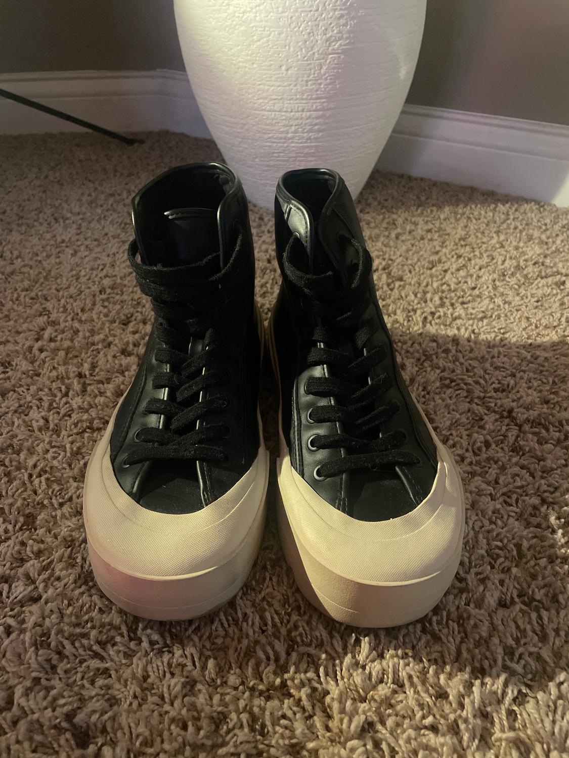 Black high-top trainers with white soles on a beige rug, next to a white lamp in the background.