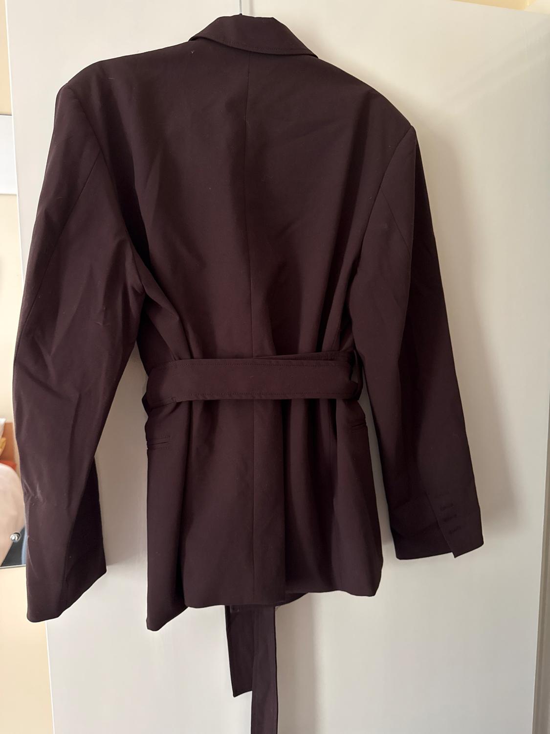 Dark brown blazer seen from behind, with a tied belt at the waist, hanging on a white door in a relaxed domestic setting.