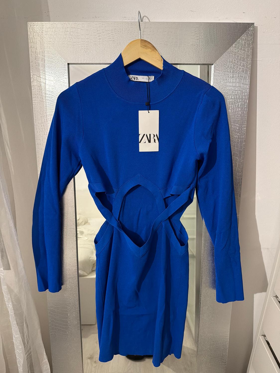 Blue dress with cut-out design at the waist, hanging on a hanger in front of a mirror. Visible label with the text "ZARA".