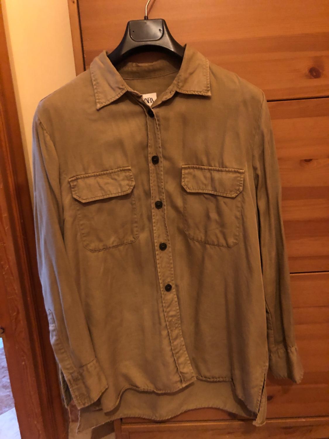 Beige long-sleeved shirt with front pockets and black buttons, hanging on a black hanger in front of a wooden wardrobe.