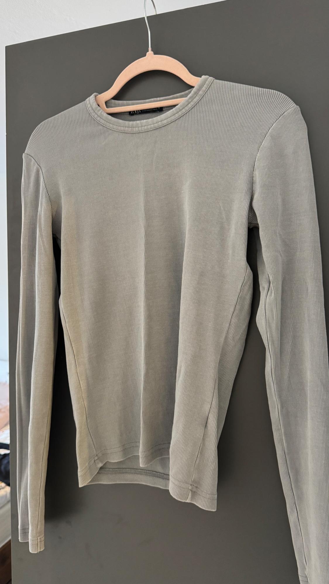 Grey long-sleeved ribbed jumper, hanging on a wooden hanger against a dark background.