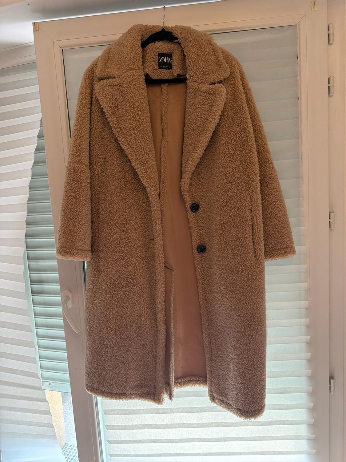 Camel-coloured coat with a bouclé texture and dark buttons, hanging in front of a window with white blinds. Inner label with the text ZARA.
