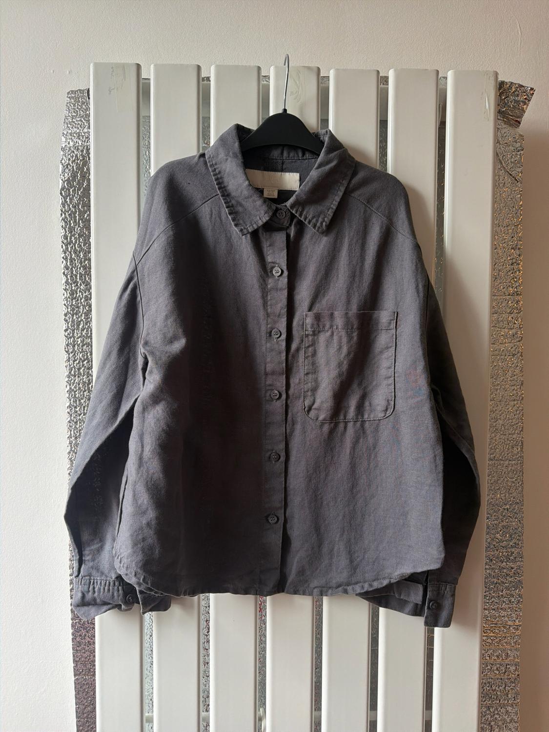 Dark grey long-sleeved button-up shirt with a front pocket, hanging on a black hanger in front of a white radiator.