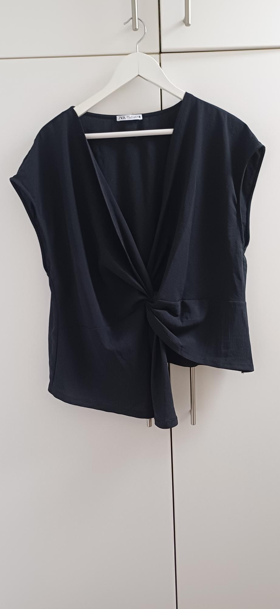 Black blouse with front knot hanging on a white hanger in front of a white wardrobe. Label visible on the collar.