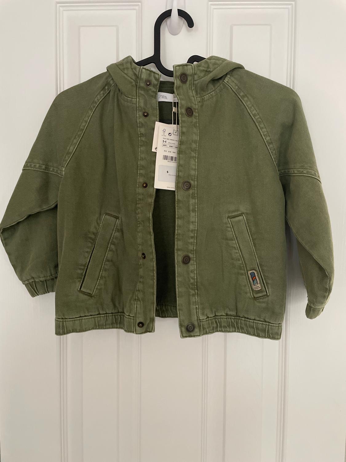 Olive green jacket with a hood and buttons, hanging on a black hanger. Visible tag with size and price details.