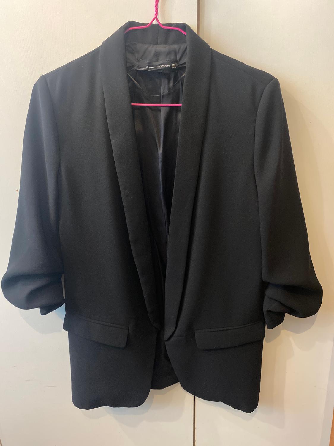 Black blazer hanging on a pink hanger, with wide lapels and rolled-up sleeves. Label visible on the inside. White background.