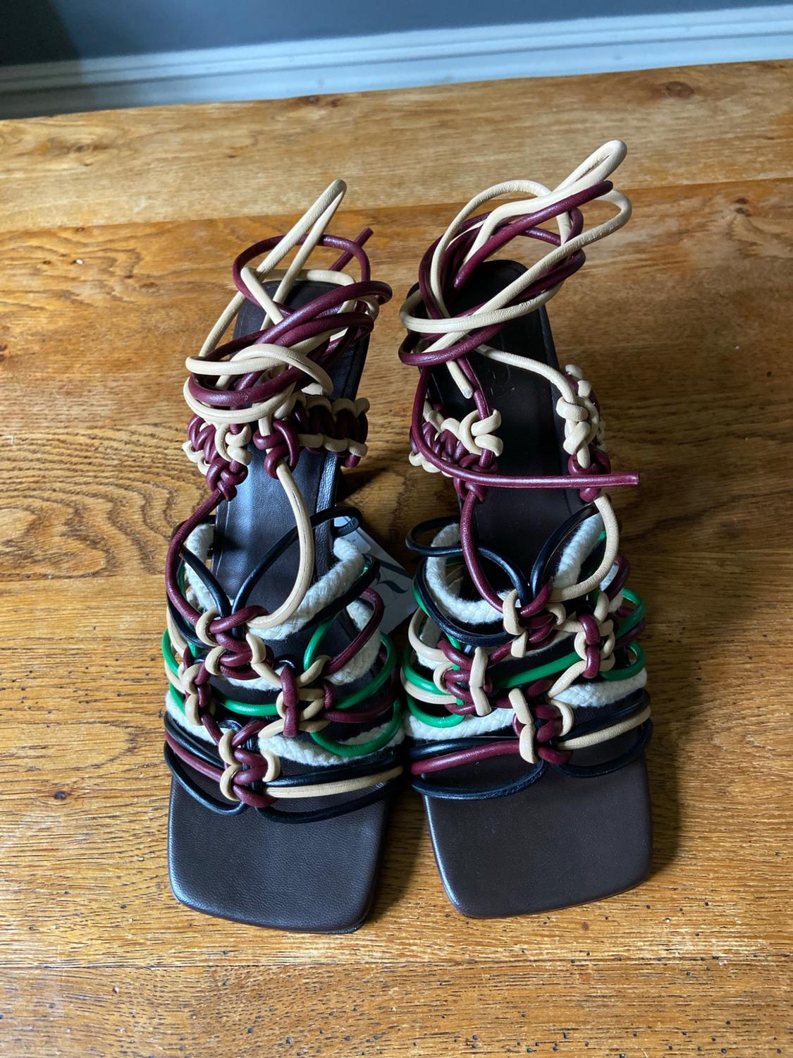 Square-toe heeled sandals with intertwined tubular straps in burgundy, beige, black, and green on a wooden floor.