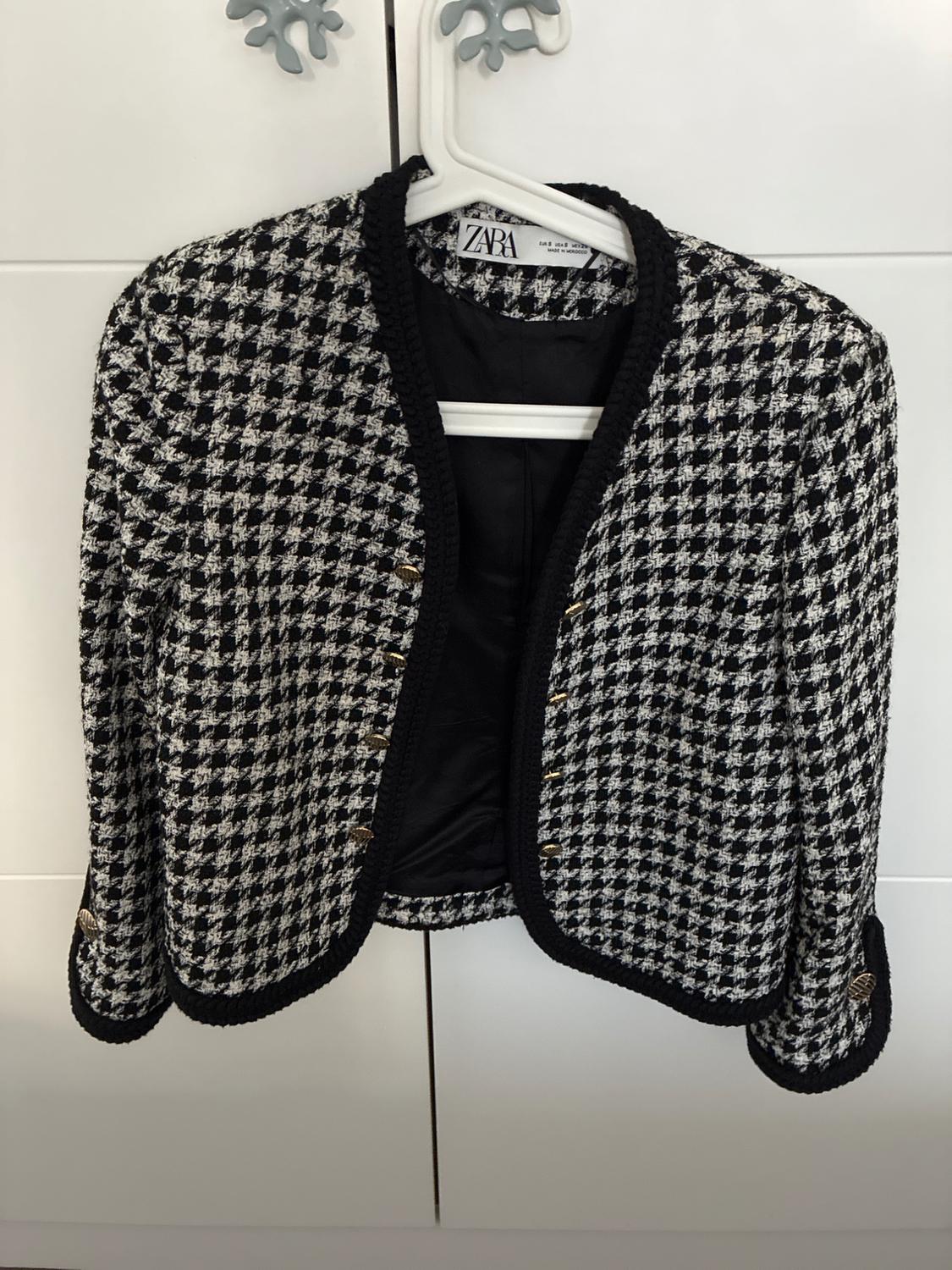 Black and white houndstooth jacket with black piping and gold buttons, hanging on a hanger. Label with the text ZARA.