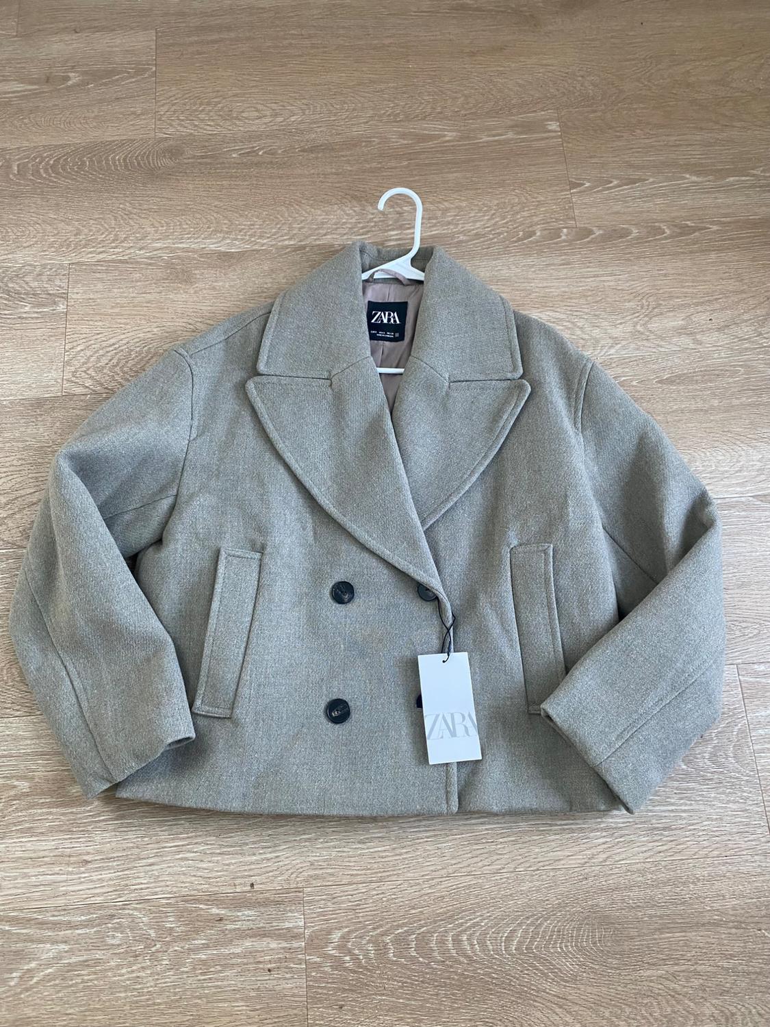 Gray double-breasted coat with wide lapels, hanging on a white hanger over a wooden floor. Visible label: ZARA.