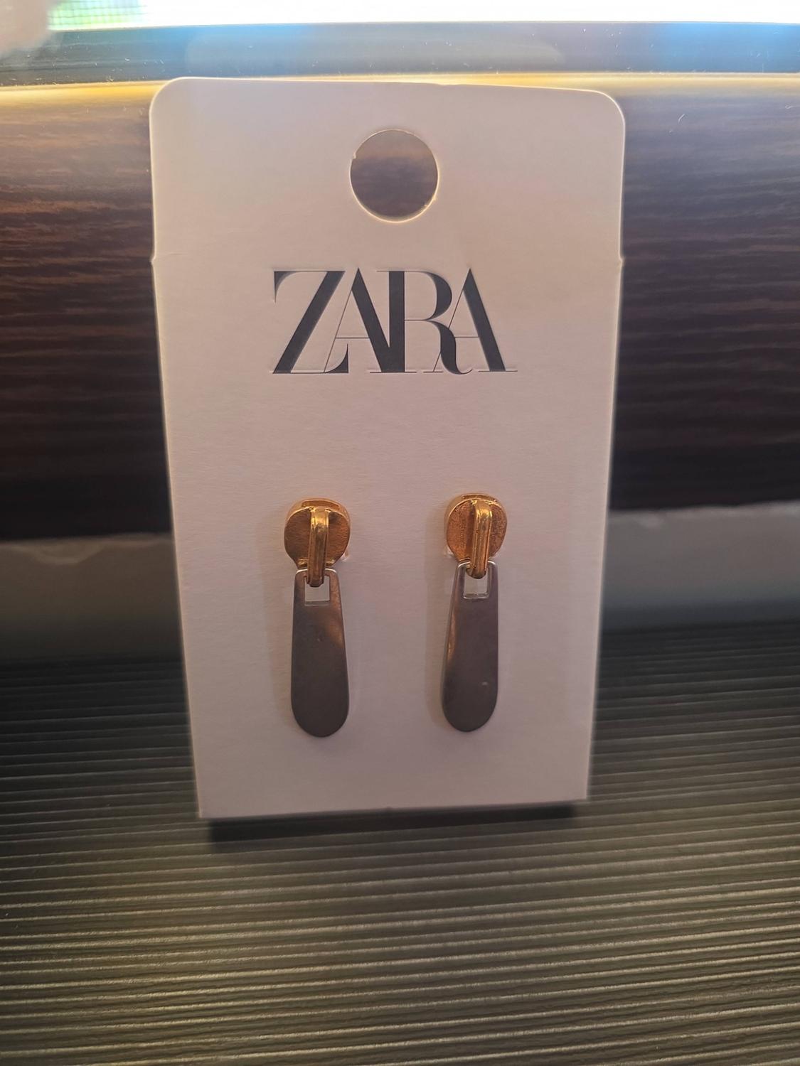 Zip-pull earrings, gold and silver, on a white tag with the text ZARA on a dark surface.