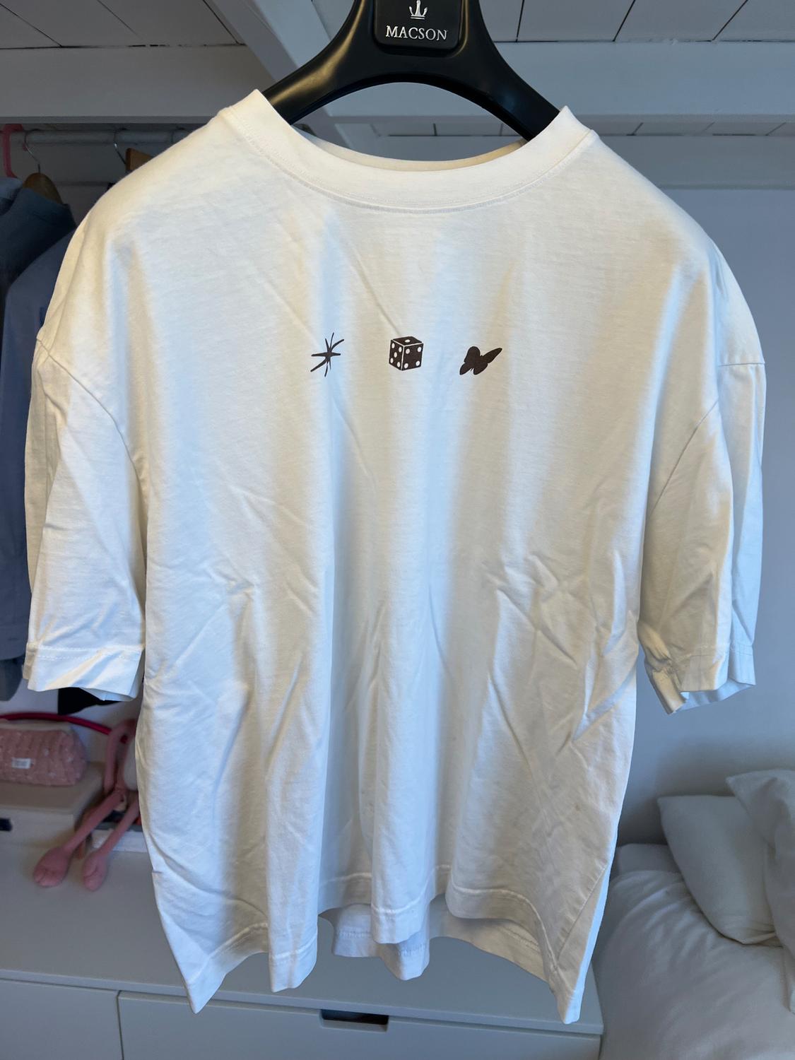 White T-shirt hanging on a black hanger with star, dice, and butterfly designs on the chest. Room background with bed and clothes.
