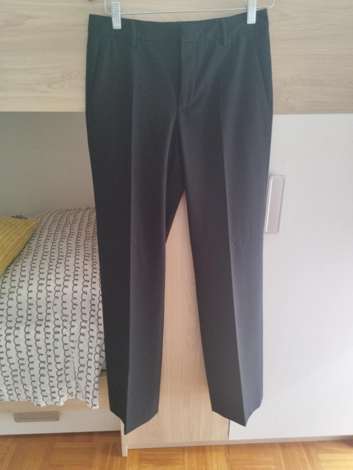 Dark smart trousers hanging on a hanger in front of a wooden wardrobe. To the left, part of a bed is visible in a homely setting.