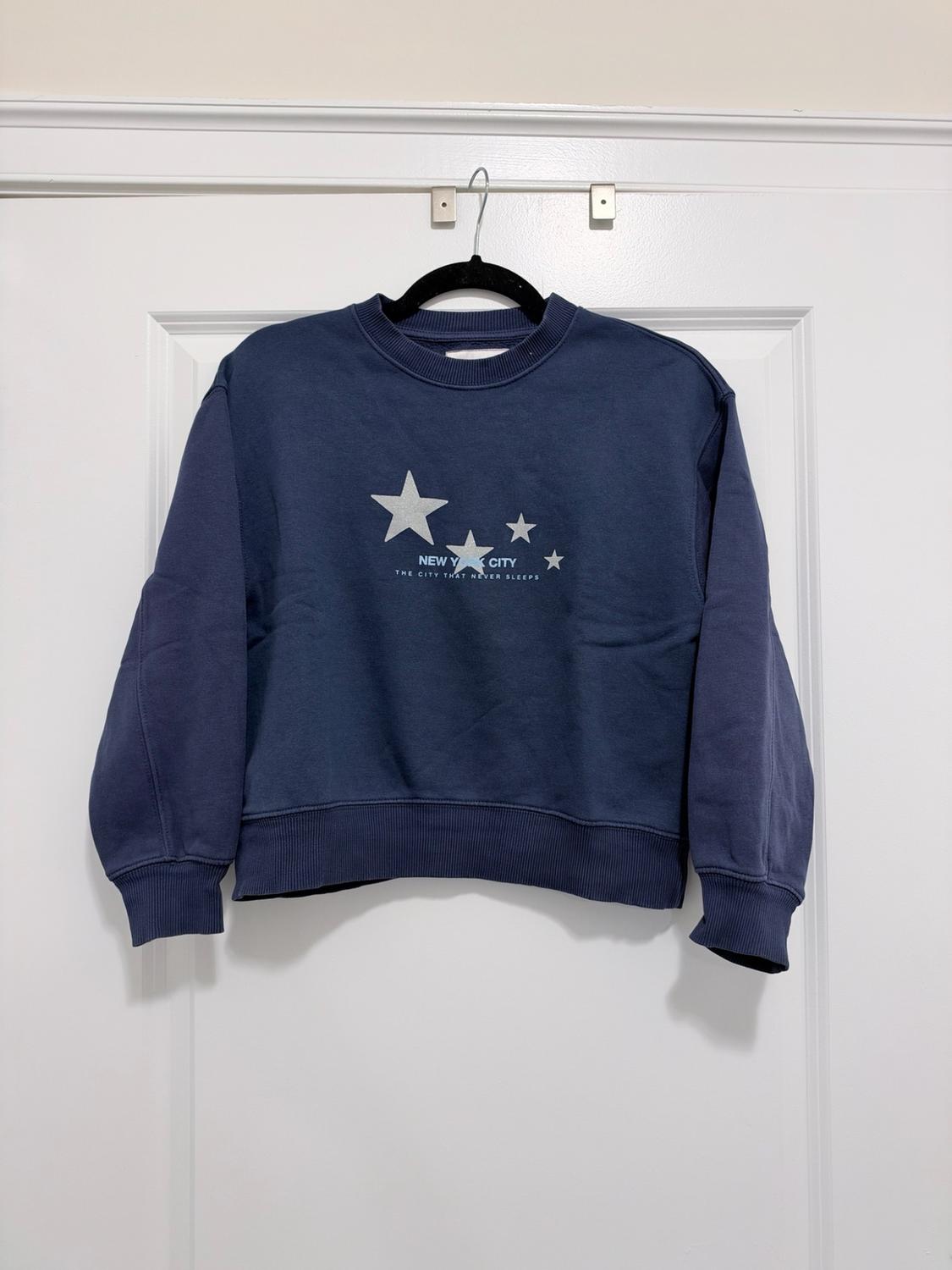 Navy blue sweatshirt hanging on a white door. Front with stars and the text NEW YORK CITY THE CITY THAT NEVER SLEEPS.