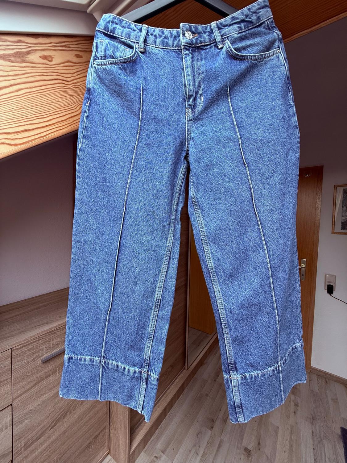 Μπλε τζιν με μπροστινές ραφές και στενή μπατζάκια, κρεμασμένα σε μια κρεμάστρα σε ένα δωμάτιο με ξύλινη κεκλιμένη οροφή.