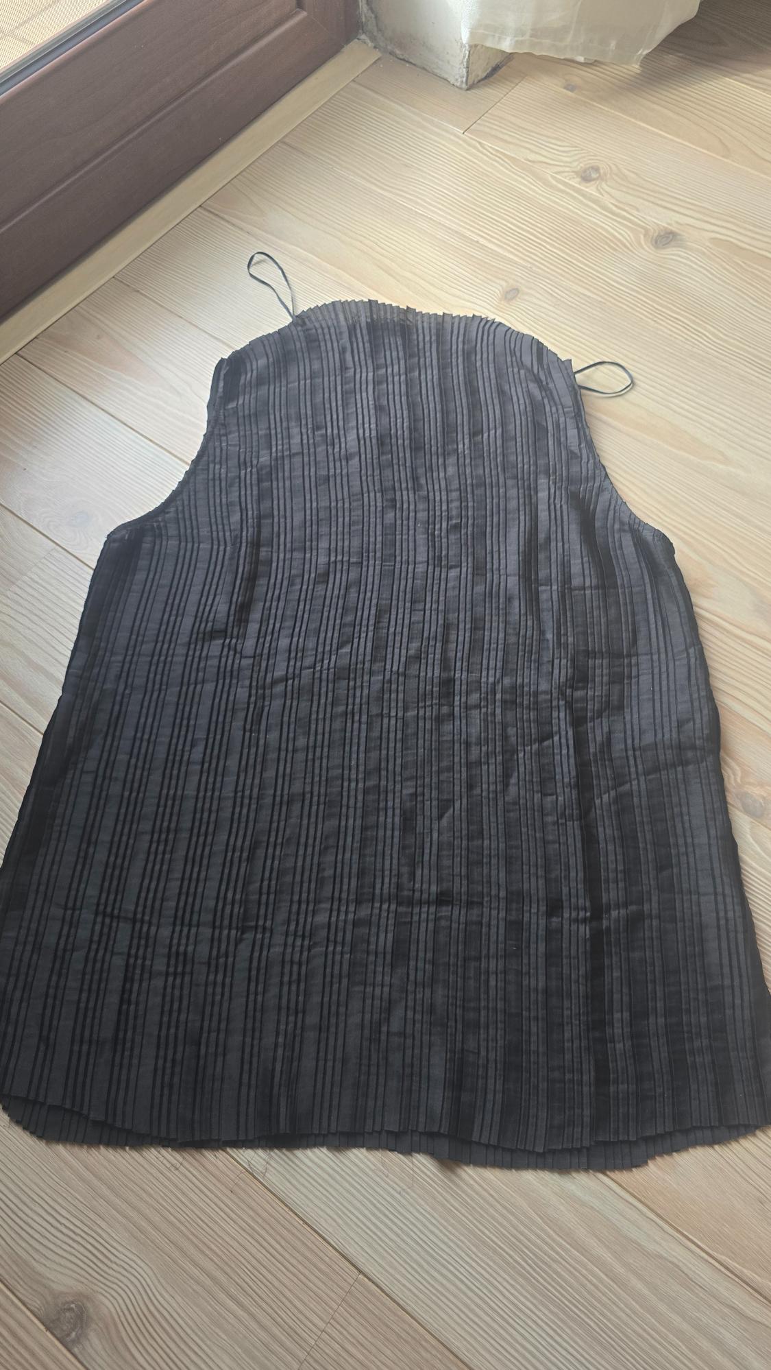 Black pleated top with thin straps, placed on a light wooden floor next to a wooden door.