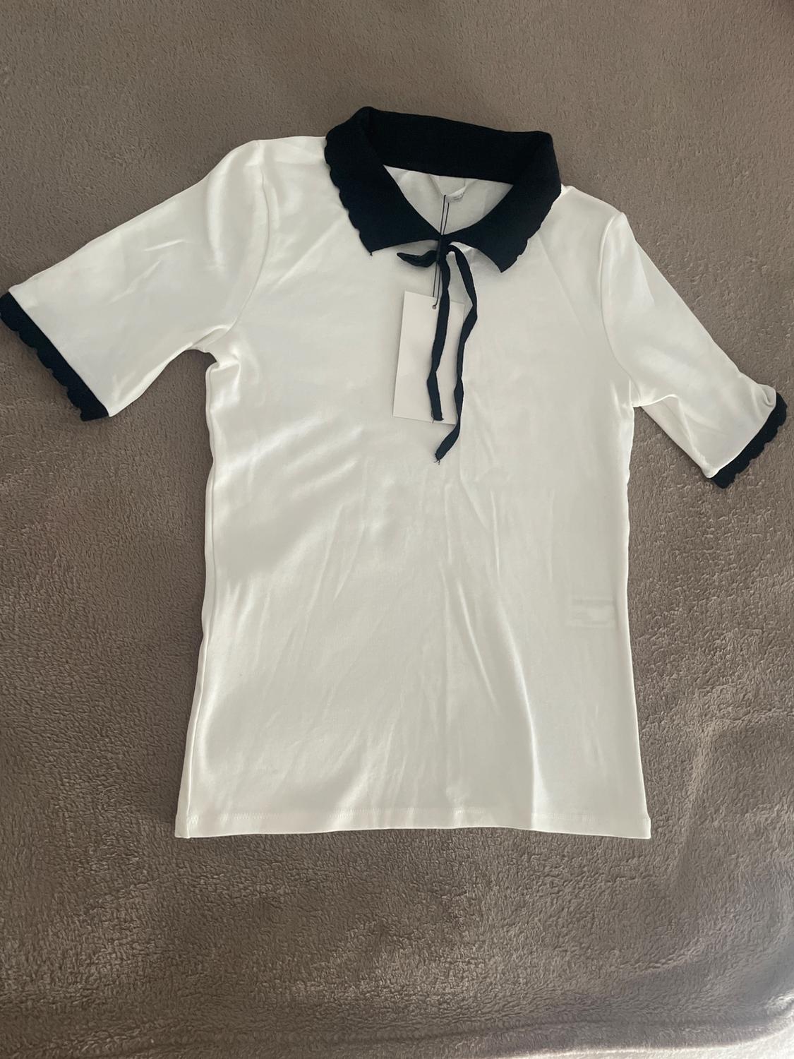 White short-sleeved T-shirt with a black scalloped collar, front tie, and black piping, set against a textured brown fabric background.