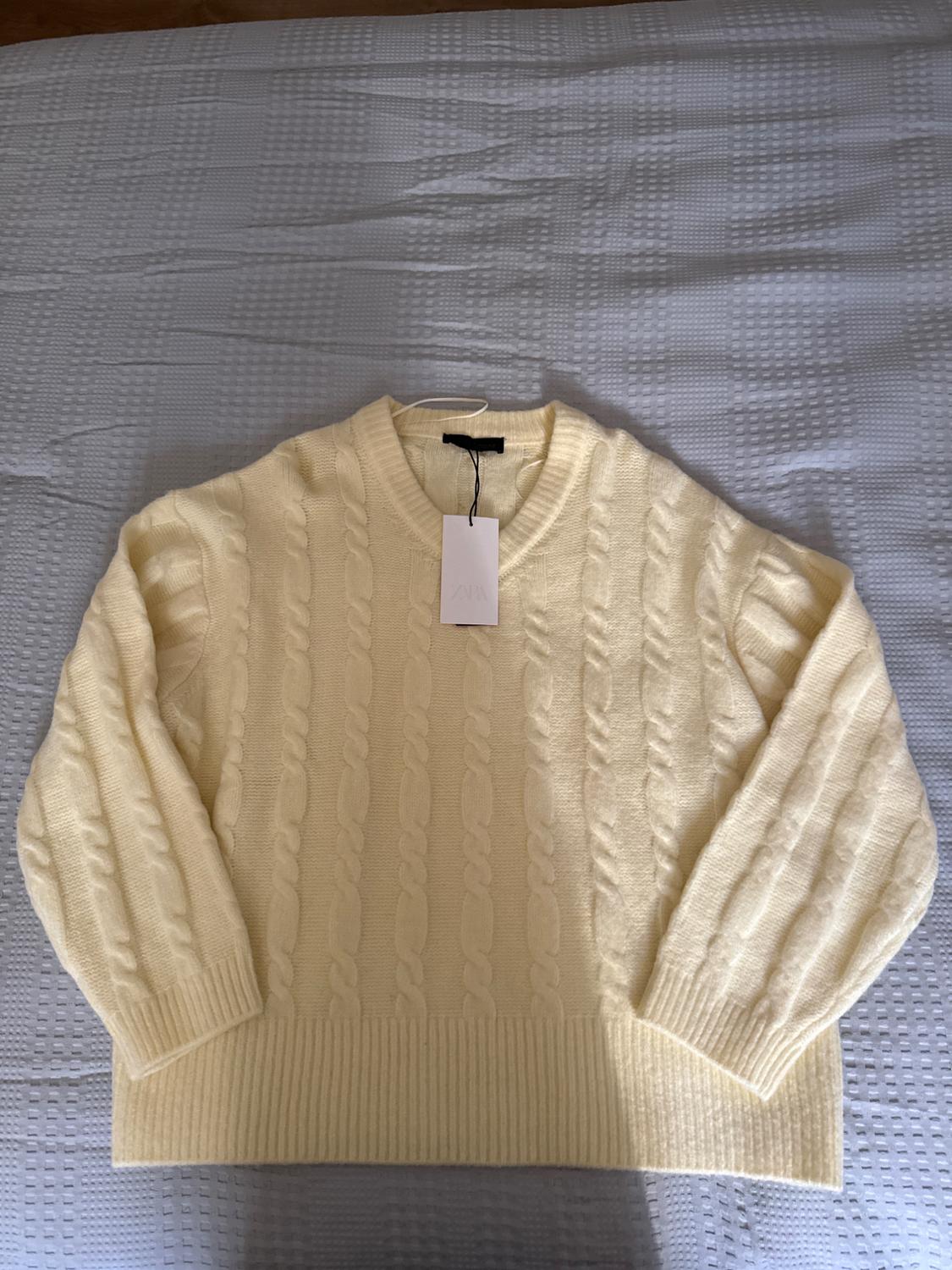 Light yellow cable-knit V-neck jumper with ZARA label, laid out on a bed with a grey bedspread. Relaxed atmosphere.