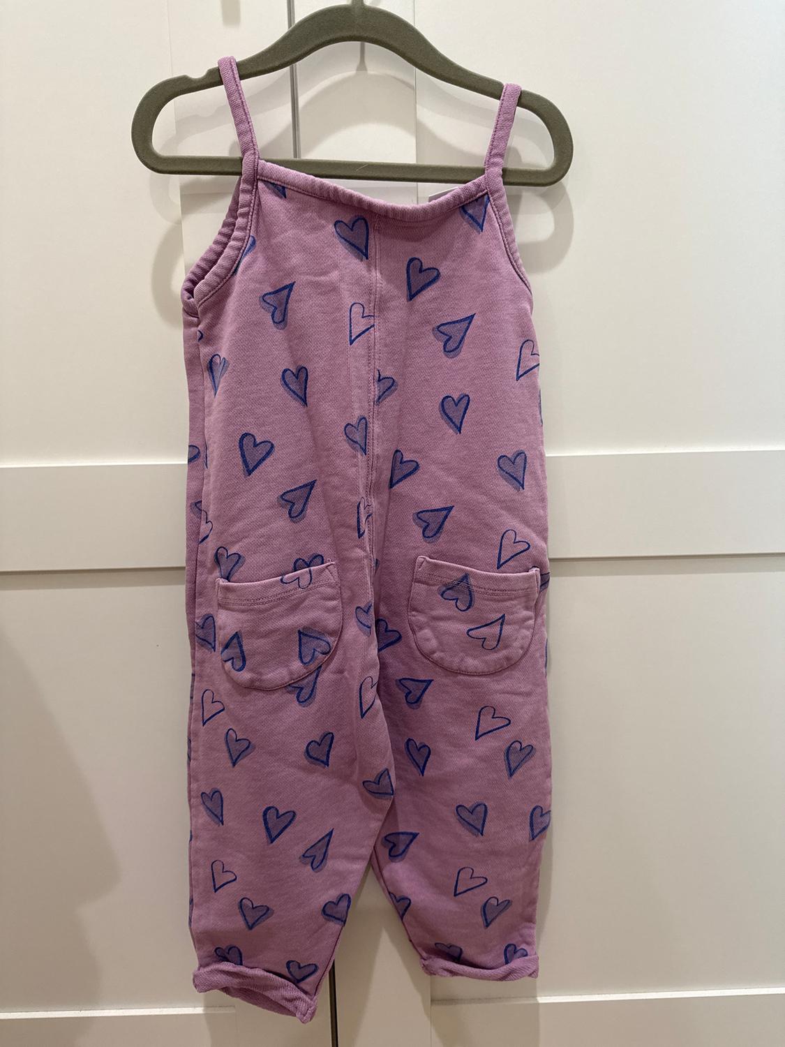 Pink children's dungarees with straps, blue heart print and front pockets, hanging on a grey hanger.