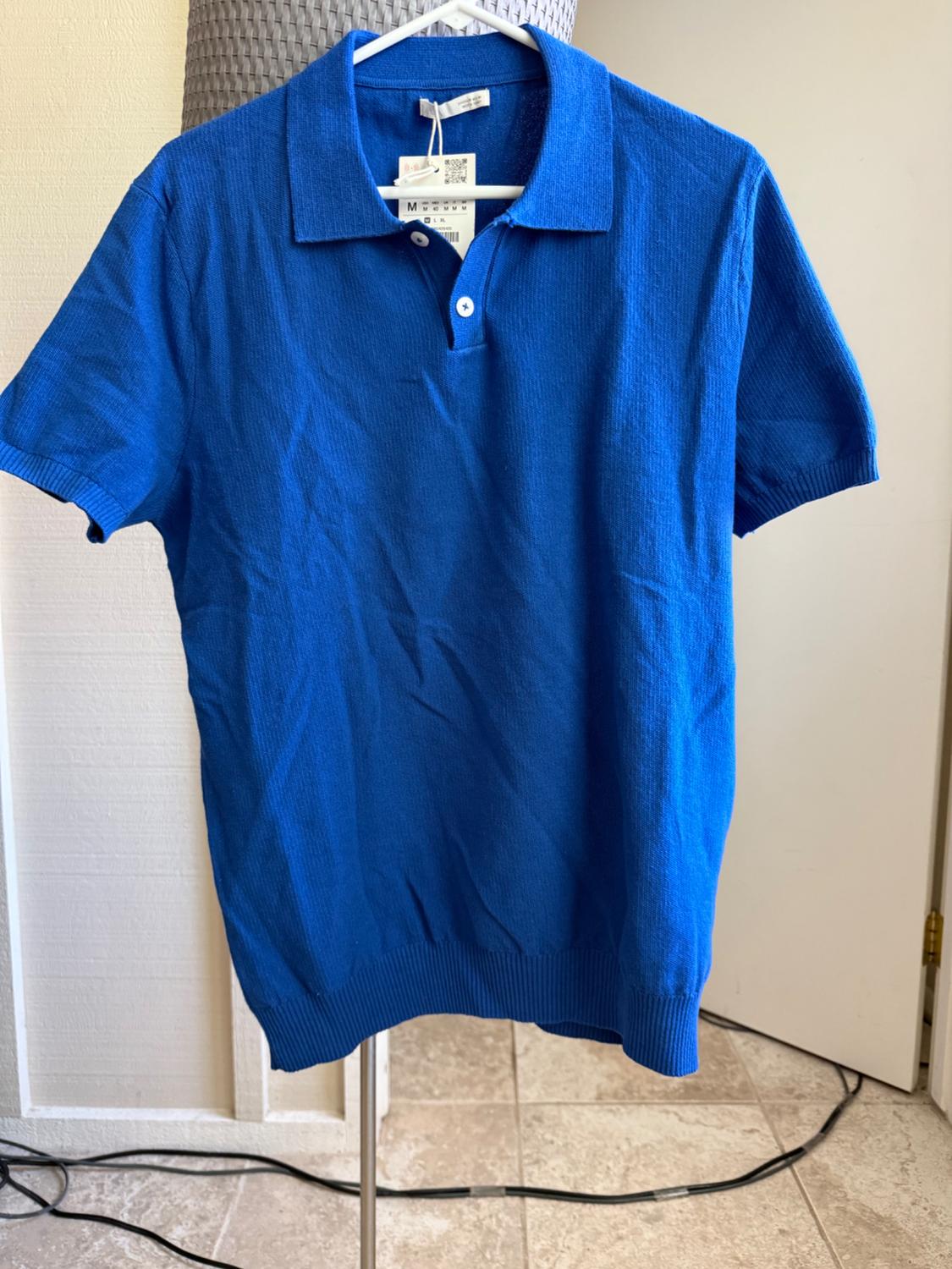 Blue short-sleeved knitted polo shirt hanging on a white hanger indoors. It has a buttoned collar and a label marked with the letter M.