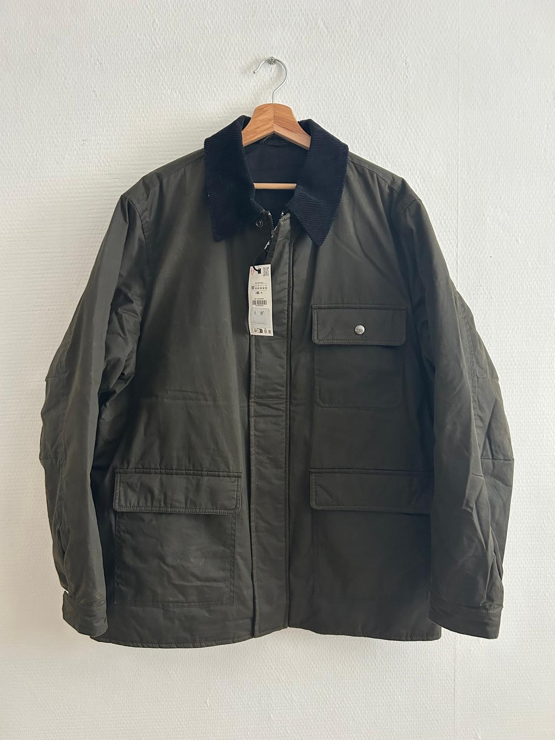 Olive green jacket with a black corduroy collar, hanging on a wooden hanger. Label visible on the front. White background.