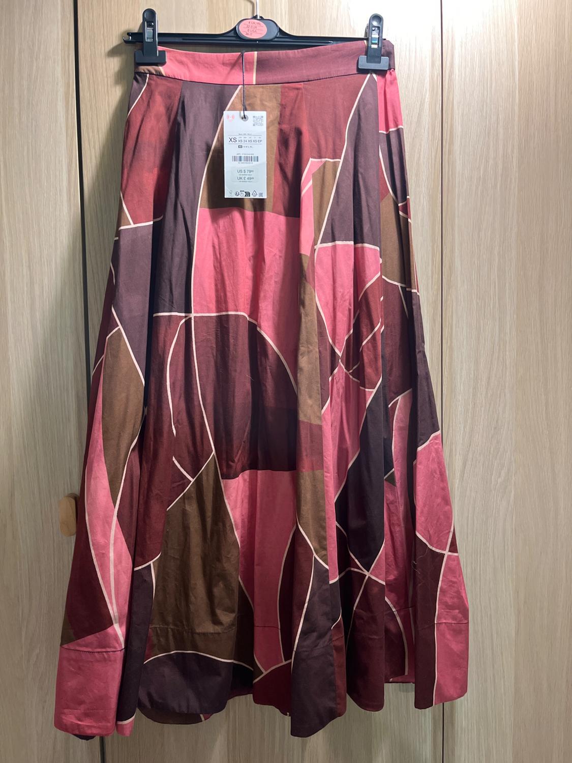 Long pleated skirt with a geometric print in pink, brown and burgundy, hanging on a hanger against a wooden background. Size XS label with text.