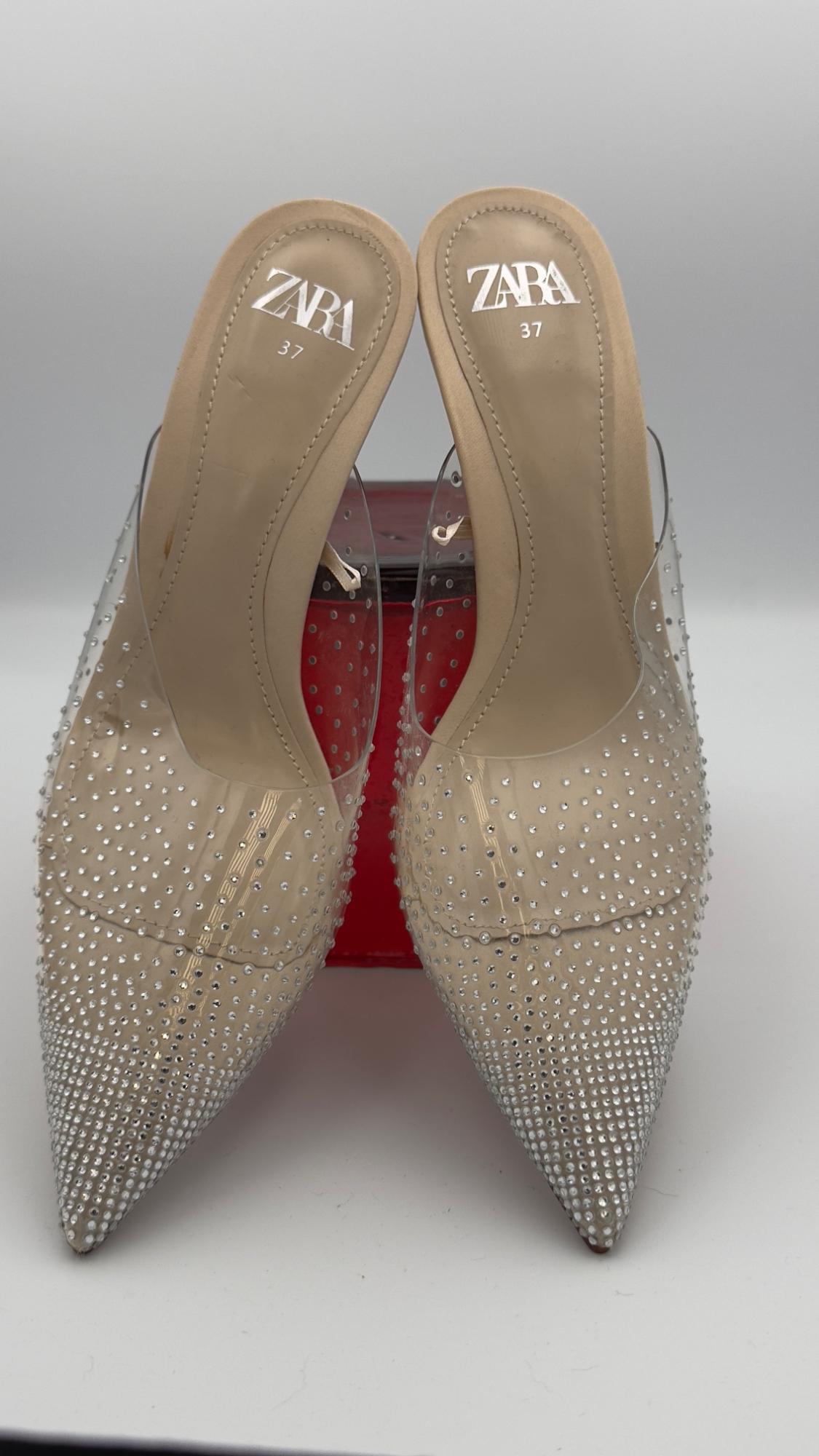 Transparent high heels with pointed toe and sparkly embellishments. Visible text: "ZARA 37". White and red background.