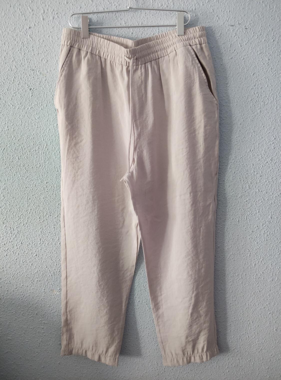 Light beige casual trousers with an elasticated waist and drawstring, hanging on a metal hanger against a light blue textured wall.