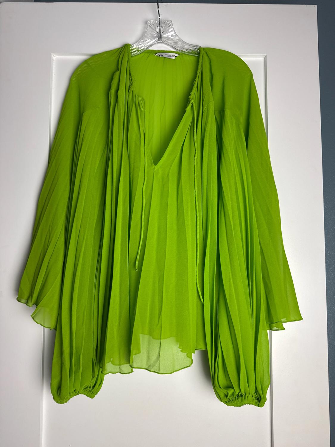 Vibrant green, pleated, semi-sheer V-neck blouse with long sleeves, hanging on a white door.