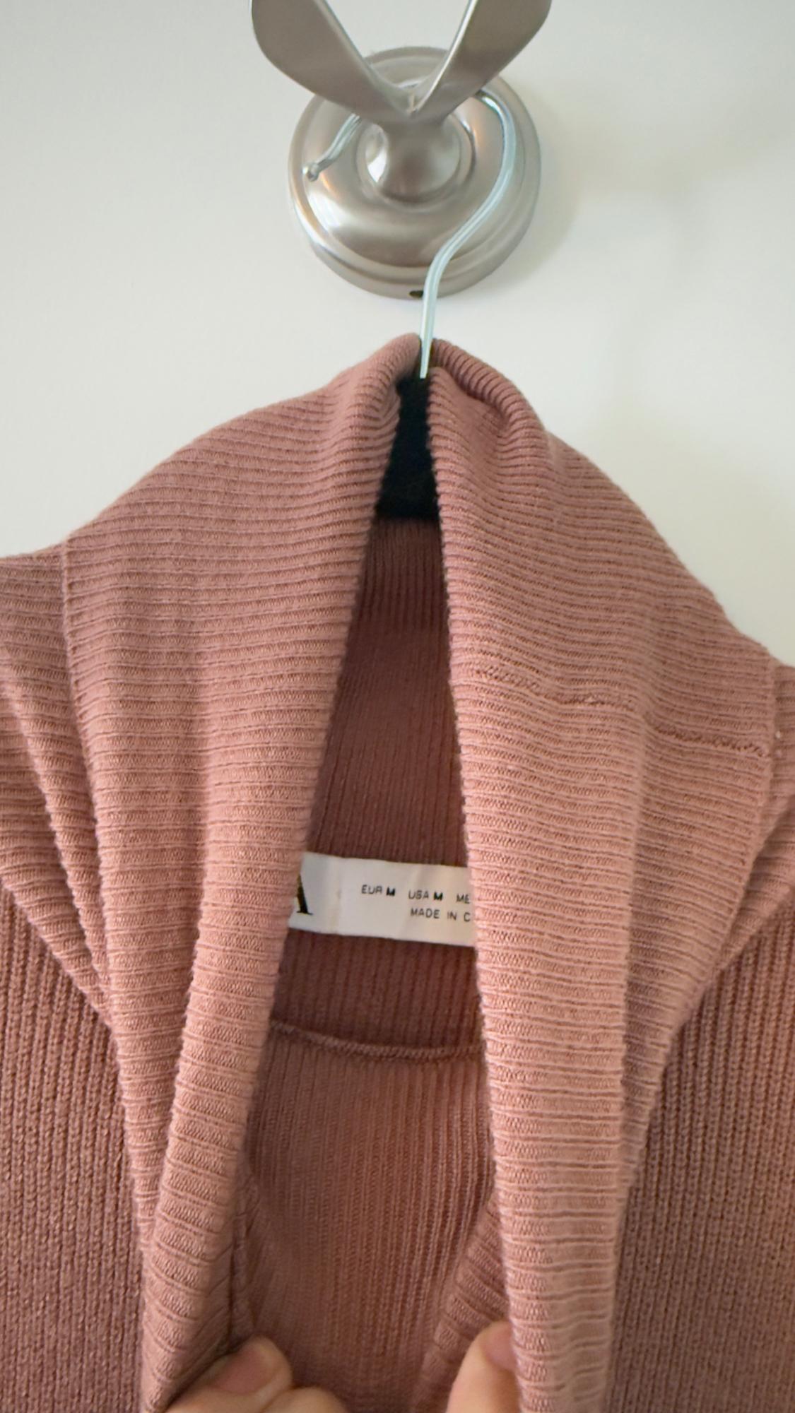 Pink knitted sweater hanging on a metal hook. Visible tag with text: "EUR M USA M MADE IN CHINA". Hands holding the collar.