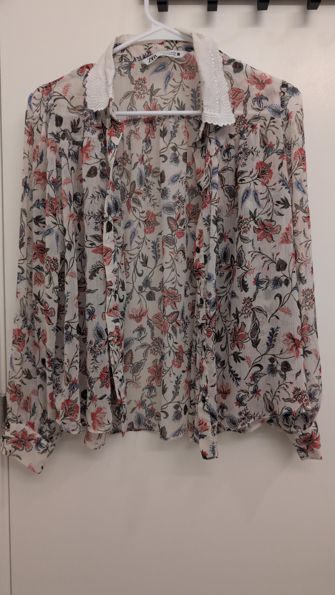 Semi-sheer blouse with a floral print in shades of red and blue, hanging on a white hanger. White collar with embroidered details.