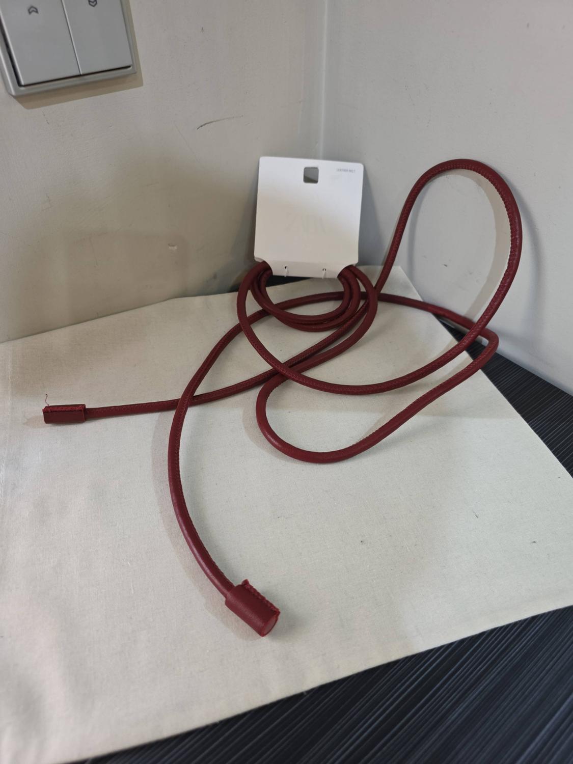 Slim dark-red cord belt with a white label, unrolled on a light-coloured fabric beside a wall.
