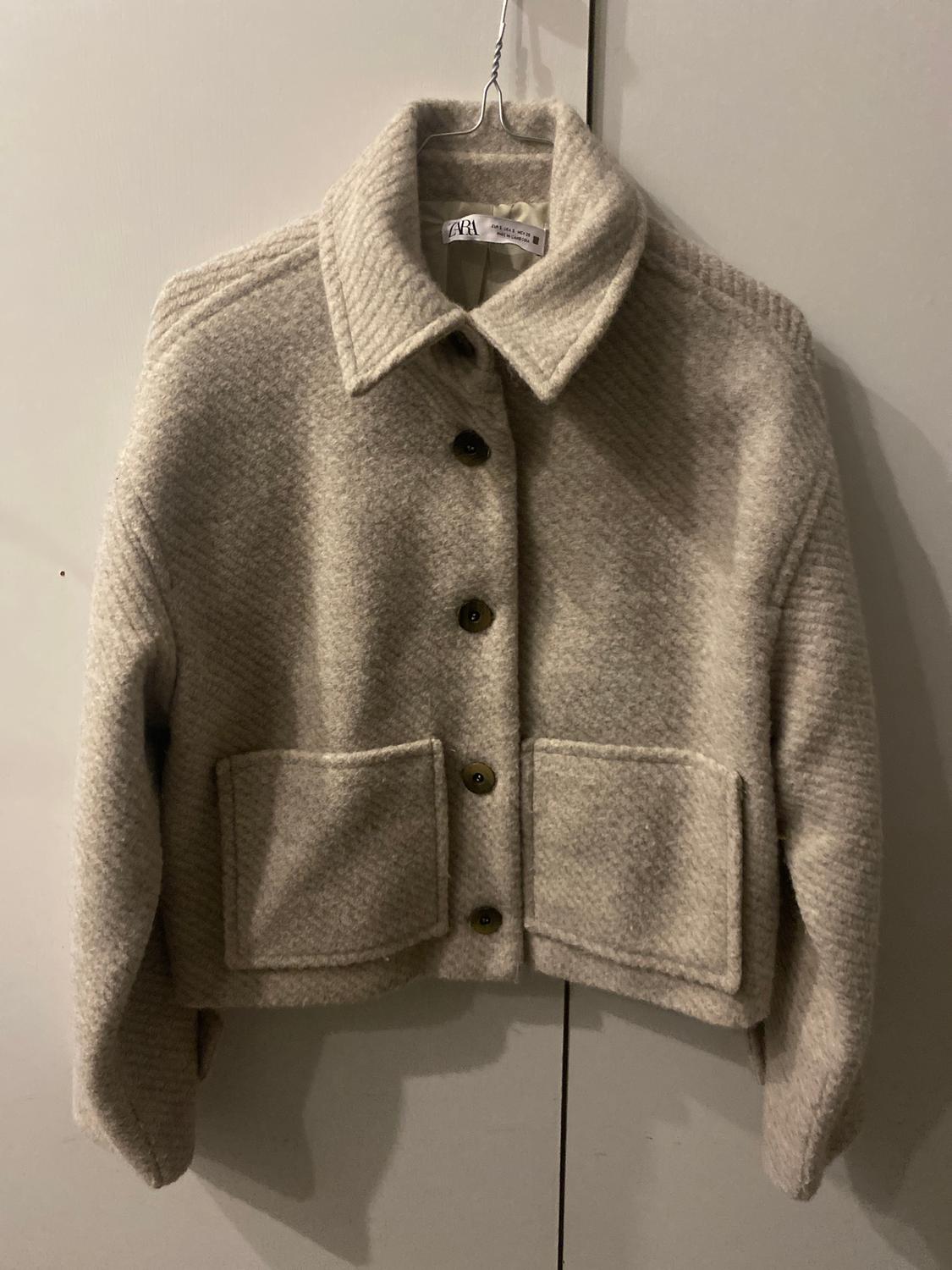 Beige wool jacket hanging on a hanger, with dark buttons and large pockets. Visible label with the brand "Zara".