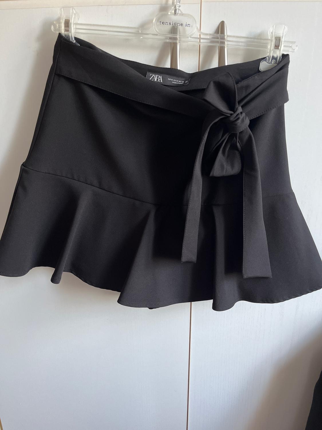 Short black skirt with a frill and bow at the waist, hanging on a clear hanger. Visible Zara tag.