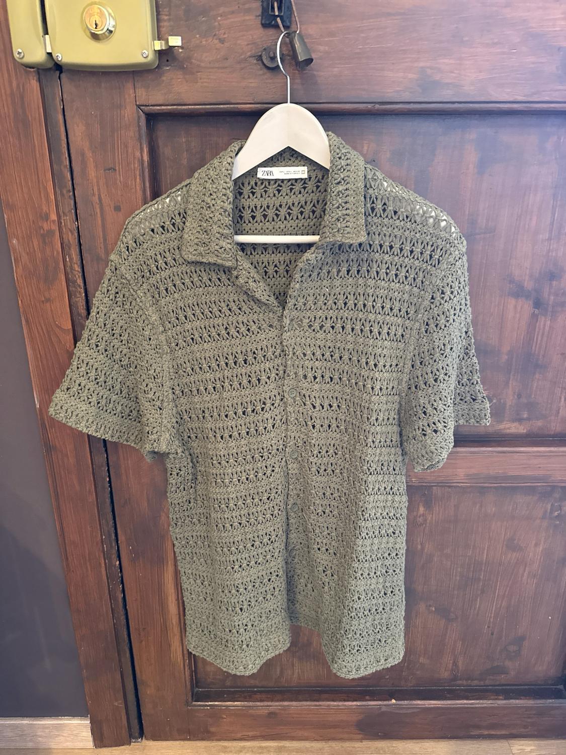 Green open-knit short-sleeved shirt hanging on a white hanger over a wooden door. The label reads ZARA.