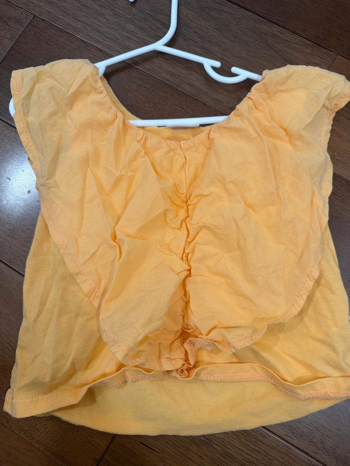 Yellow sleeveless blouse with ruffles, hanging on a white hanger over a brown wooden floor.