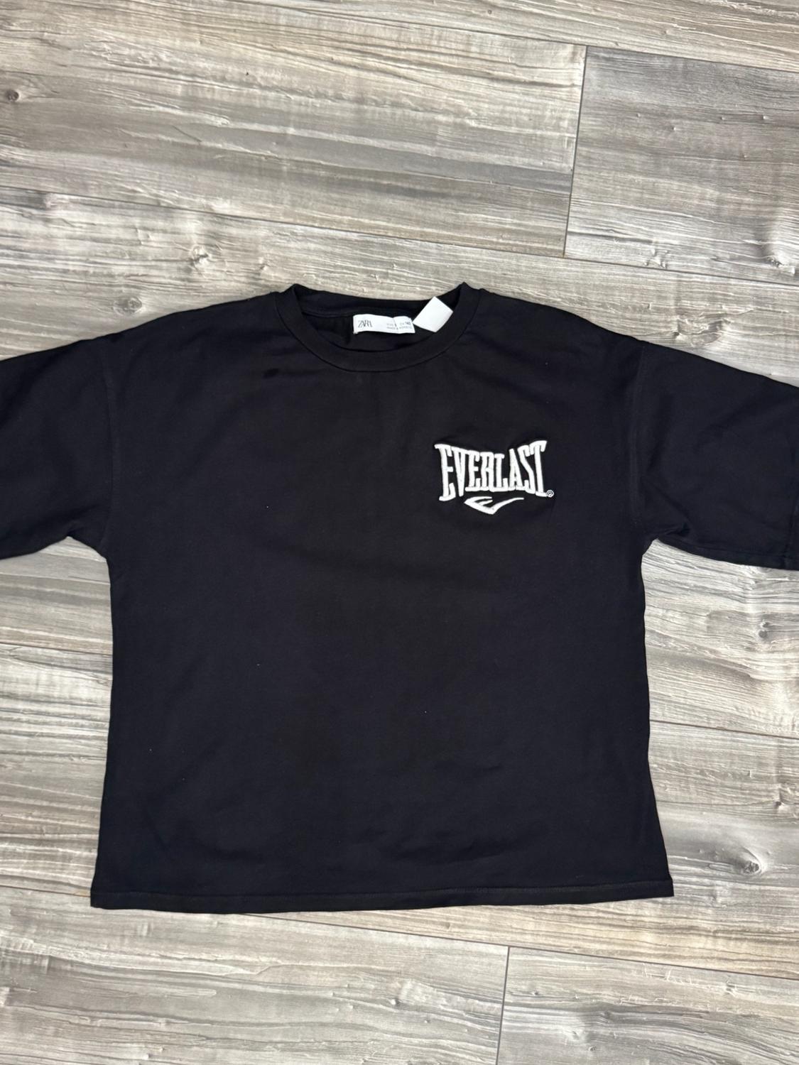 Black short-sleeved T-shirt with the text EVERLAST printed in white on the chest, laid out on a wooden floor.