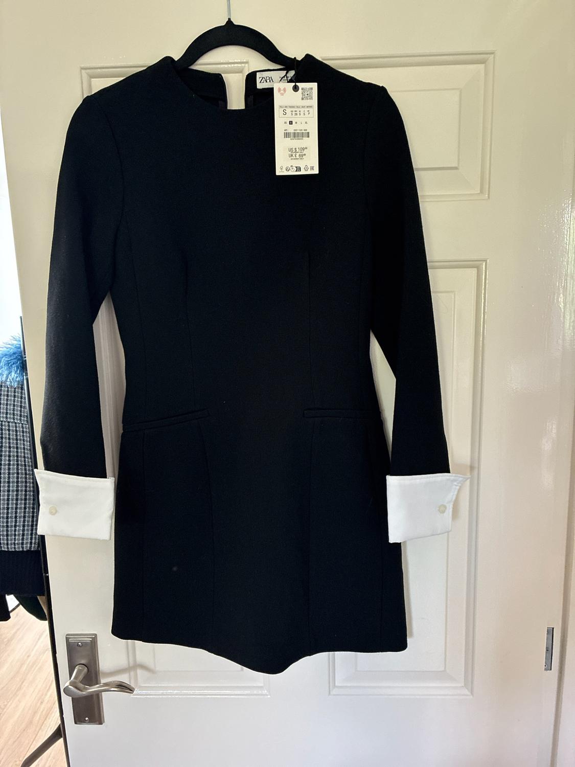Black short-sleeved long-sleeved dress with white cuffs hanging on a white door. Label with text ZARA S US $ 109 UK £ 69.