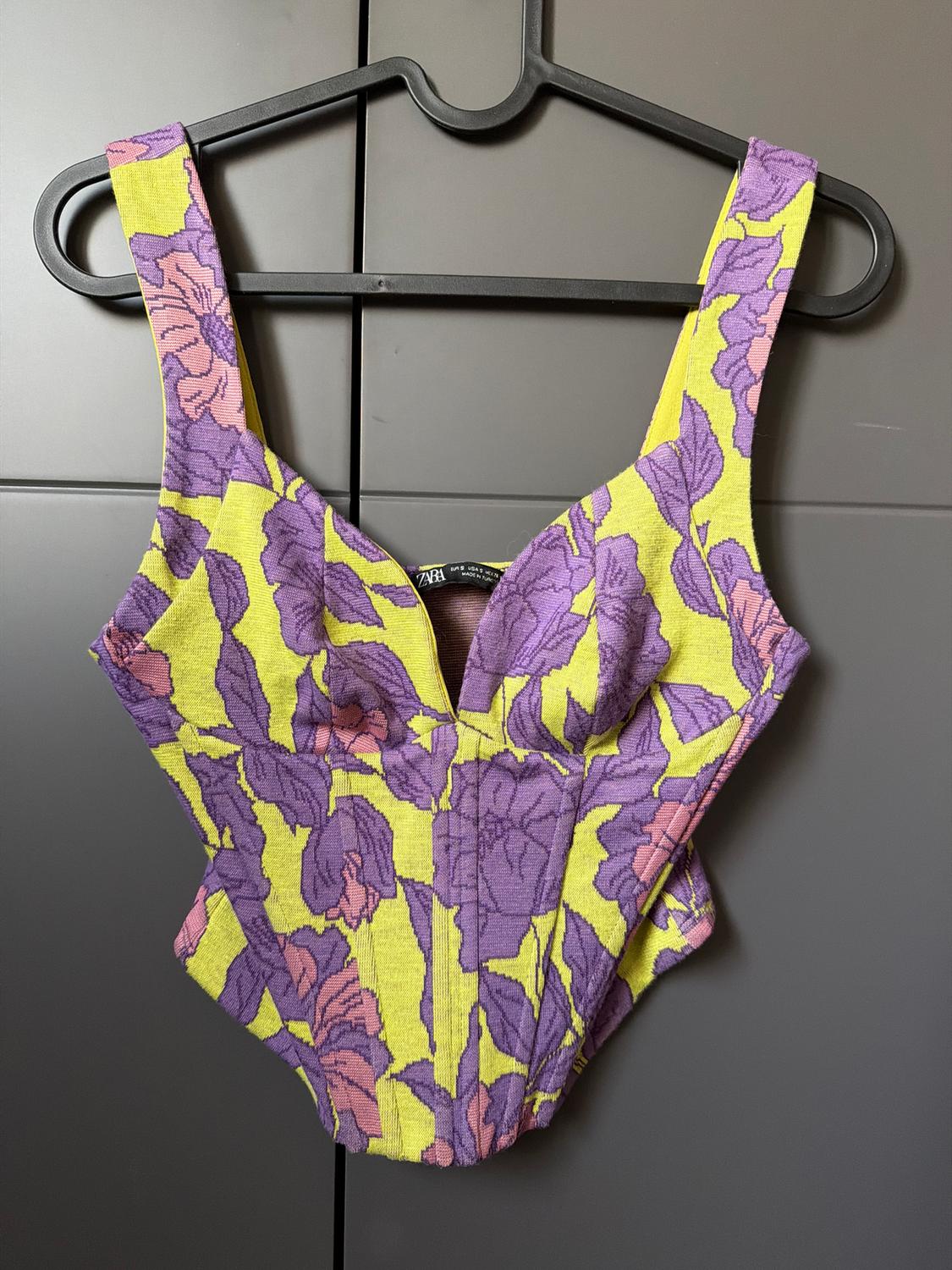 Sleeveless top with a floral print in purple and yellow tones, hanging on a black hanger against a grey background.