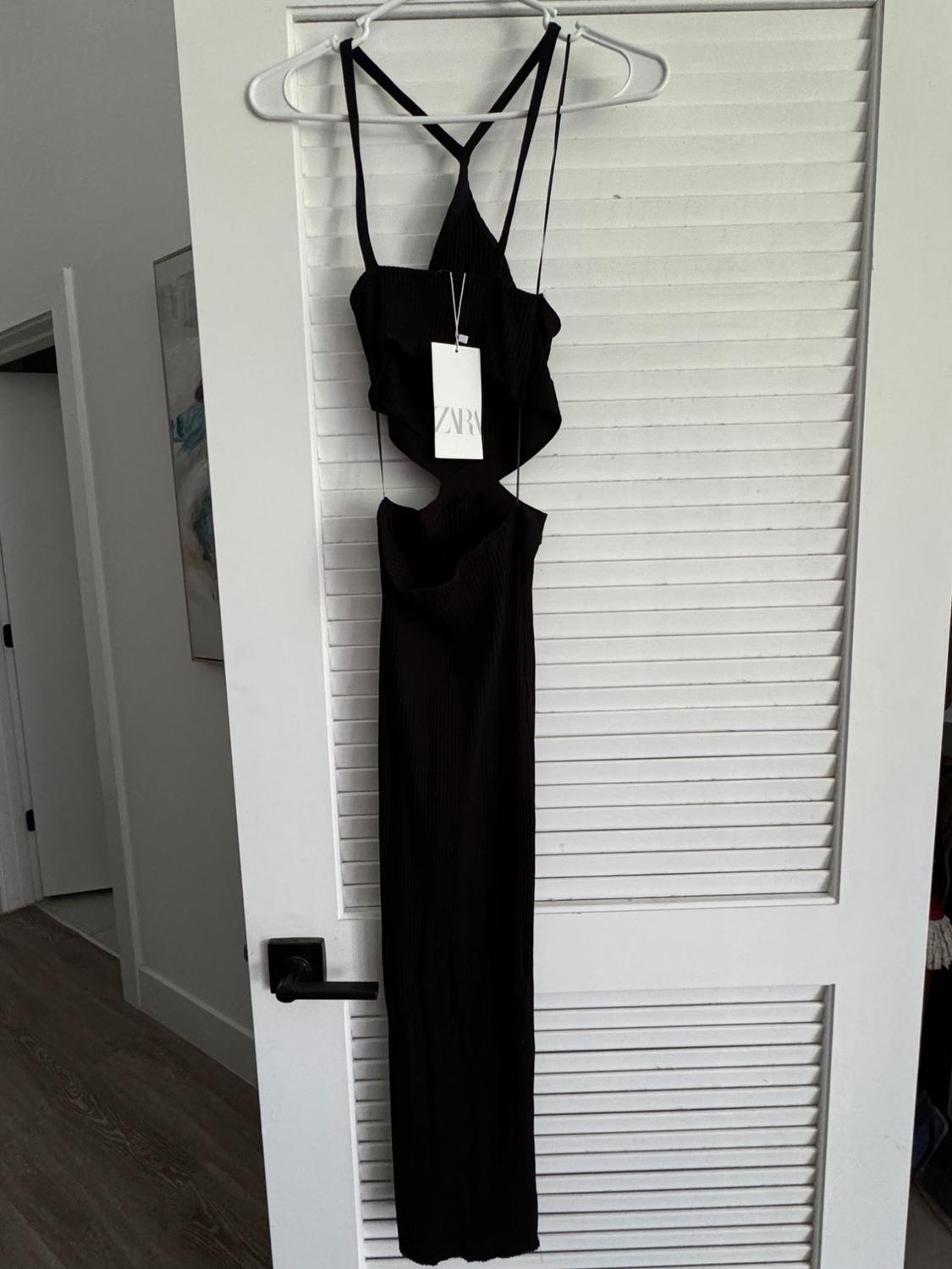 Long black dress with thin straps and a cinched waist, hanging on a white door. Visible tag with the text "ZARA".