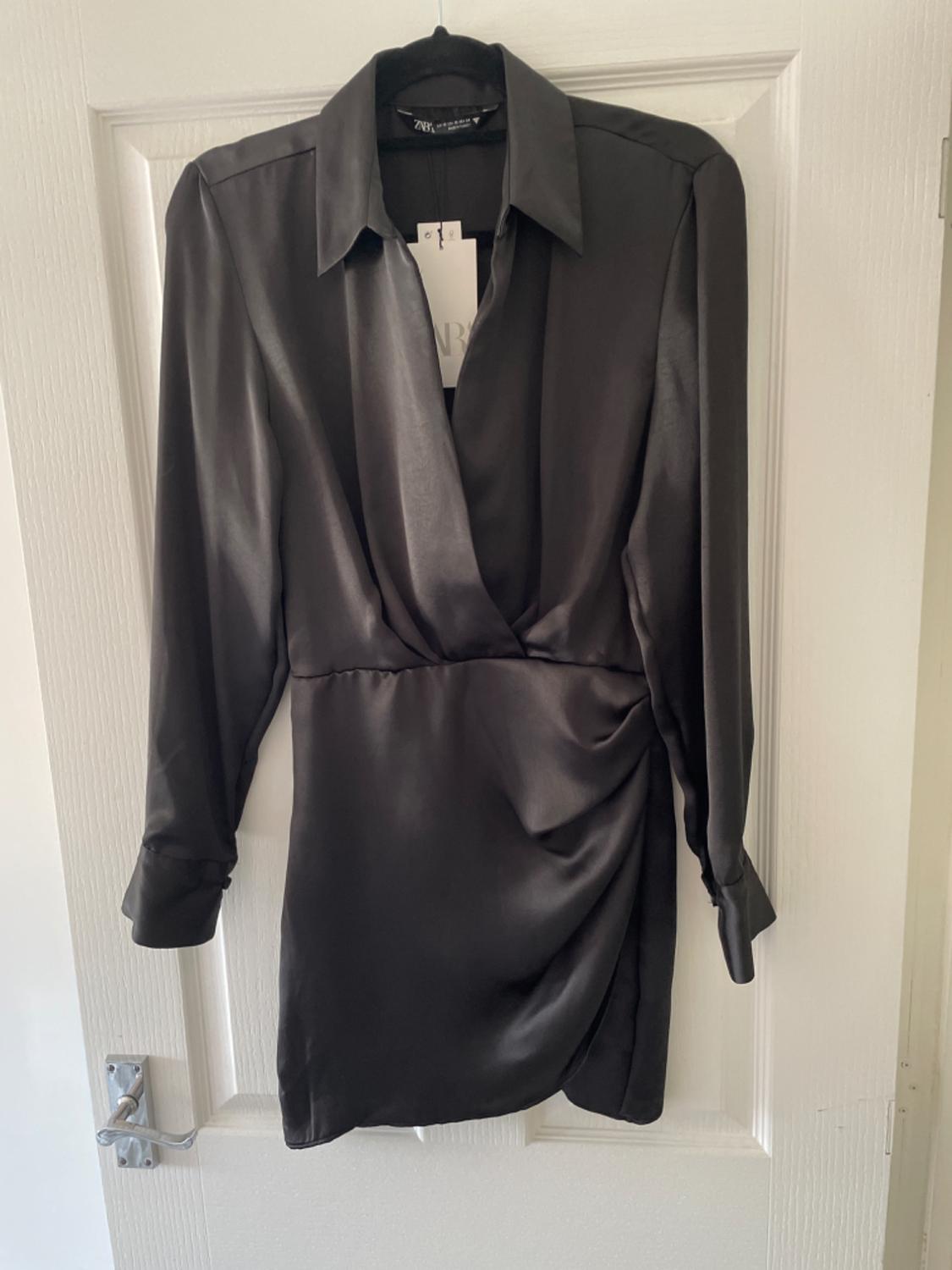 Short black satin long-sleeved dress, with a wrap neckline and draped detailing, hanging on a white door. ZARA label visible.