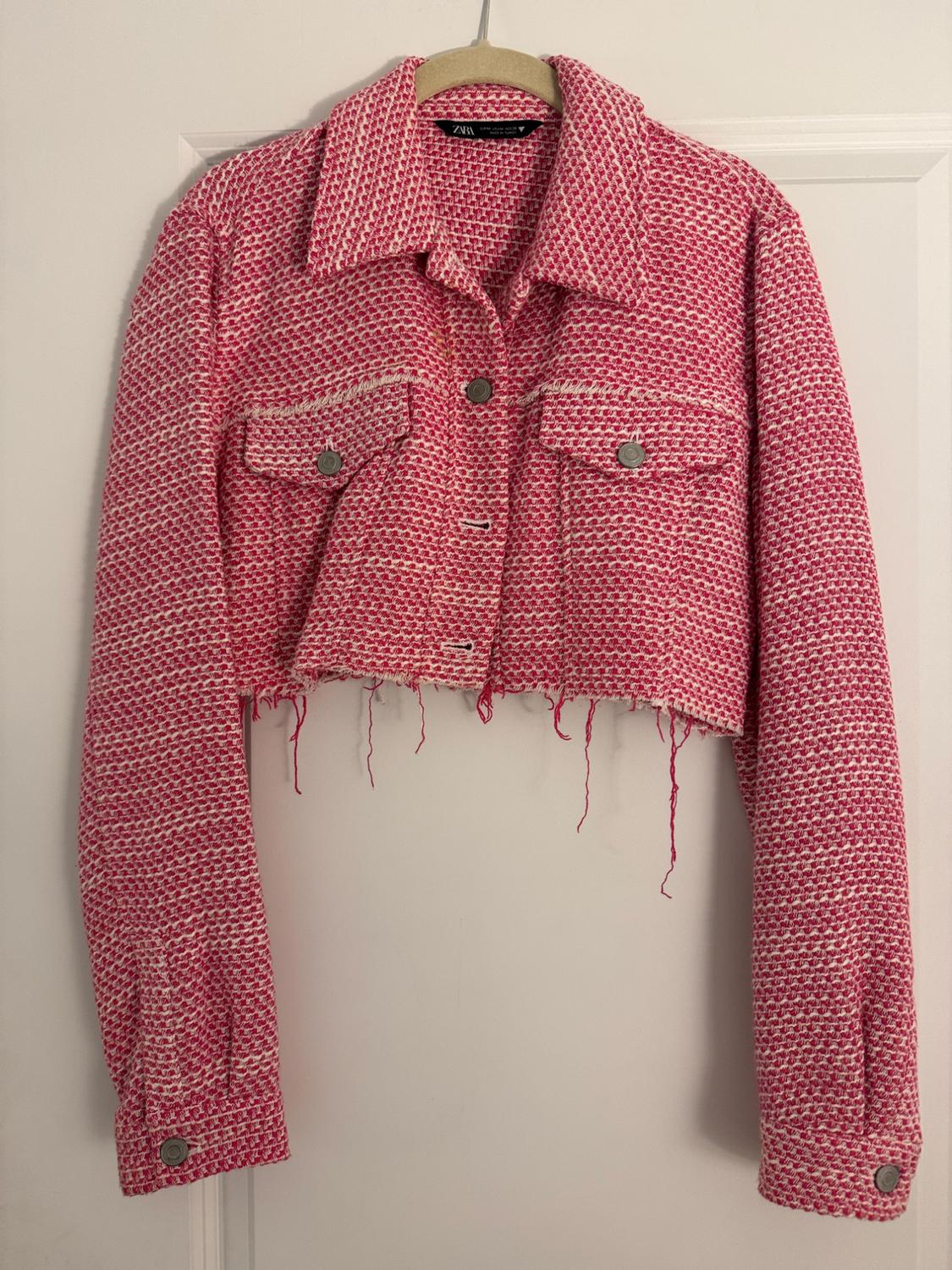 Short pink and white woven jacket with metal buttons, front pockets and frayed edge, hanging on a beige hanger.