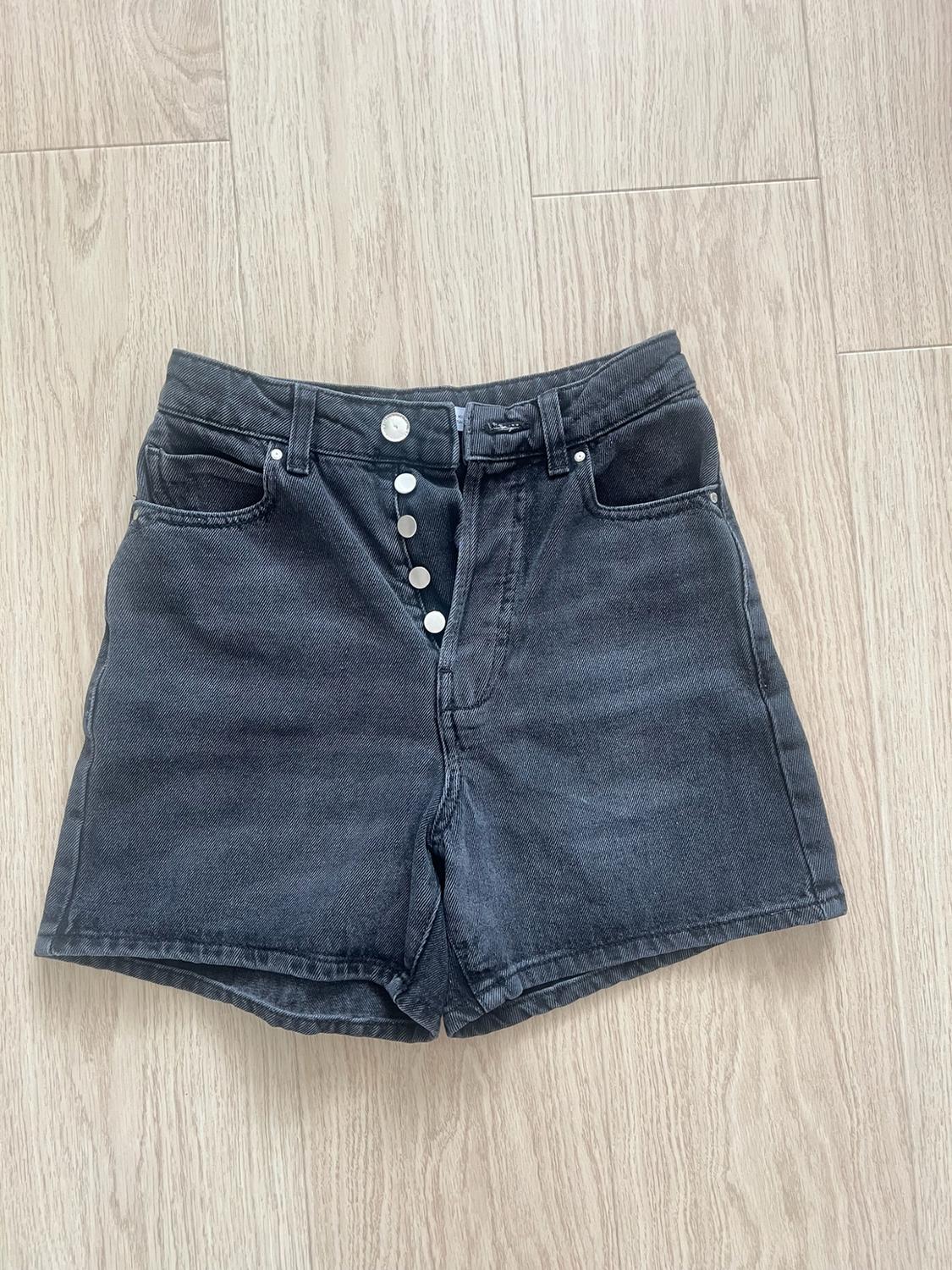 Black denim shorts with silver front buttons, photographed from above on a light wood floor in a casual style.