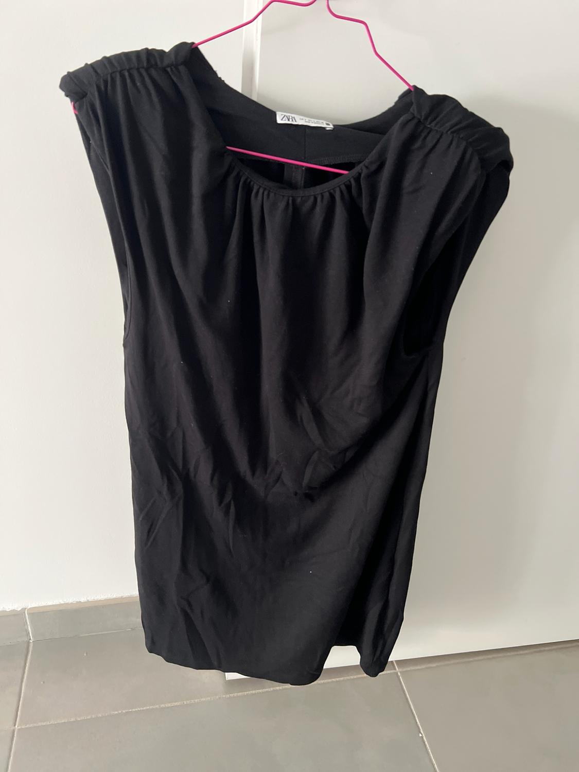 Black sleeveless top with gathered neck hanging on a pink hanger in front of a white door and grey floor. ZARA label.