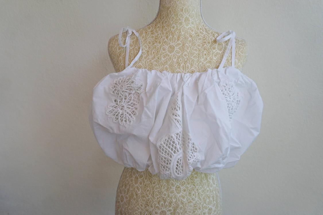 White top with lace details on a mannequin with a beige background. Design with thin straps and voluminous texture.