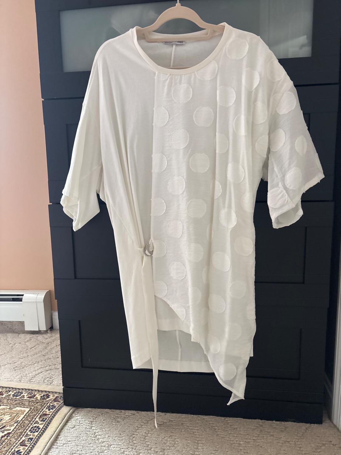 White short-sleeved t-shirt with large polka dots, hanging on a hanger in front of a black wardrobe. Room background with a rug.