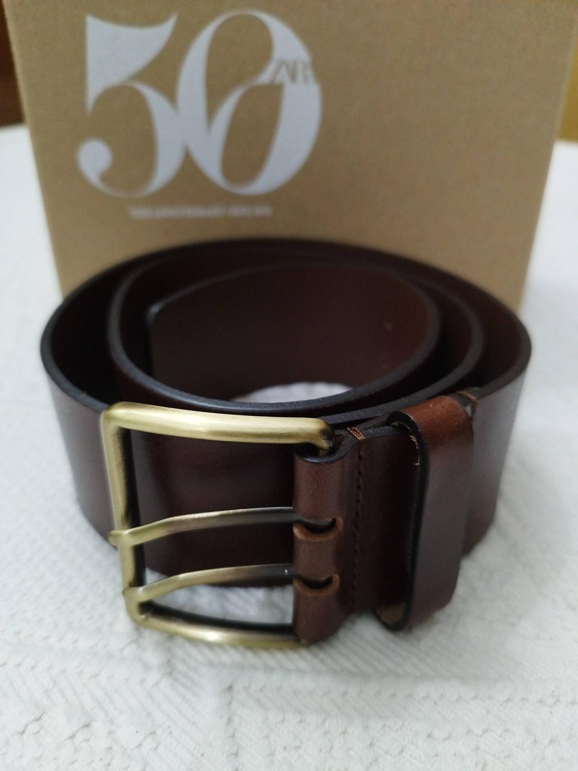 Rolled brown belt with a gold buckle on white fabric. Behind it, a cardboard box with the text 50 ZARA.