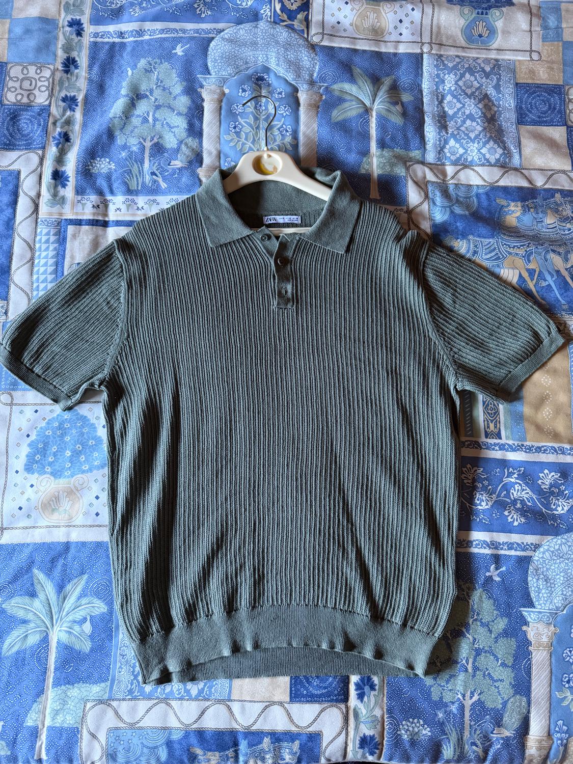 Green ribbed knit polo shirt with buttons at the neck, draped over a blue bedspread with a tree and column print.