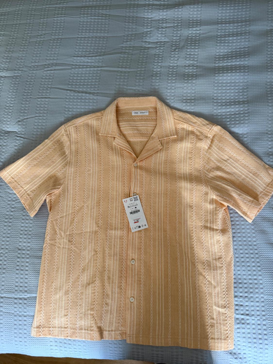 Light orange short-sleeved shirt with textured white stripes, on a blue bedspread. Labels with the text ZARA and XL.